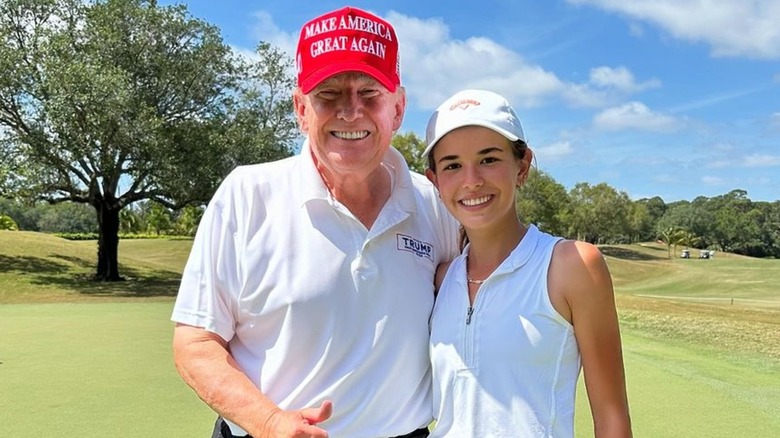 Kai Trump swinging a golf club with excellent form, while President Trump watches with a proud, smiling expression in the background. October 2025.